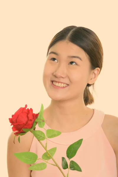 Studio ujęcie młodej szczęśliwej Azjatki uśmiechniętej i trzymającej czerwoną różę podczas myślenia — Zdjęcie stockowe