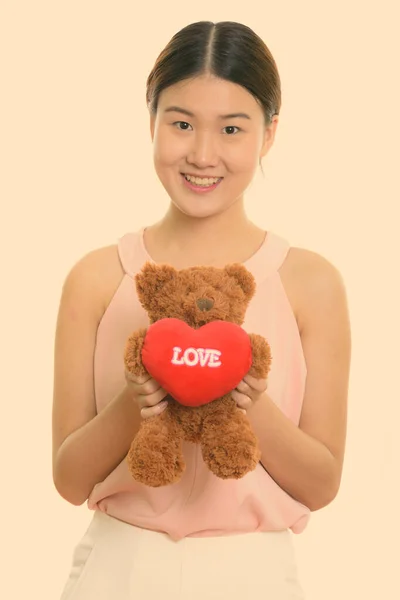 Studio shot de tineri fericit asiatic femeie zâmbind și ținând ursuleț de pluș cu inima și semn de dragoste — Fotografie, imagine de stoc