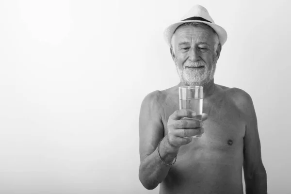 Estúdio tiro de homem barbudo sênior feliz sorrindo enquanto olha para o vidro de água e vestindo chapéu sem camisa contra fundo branco — Fotografia de Stock