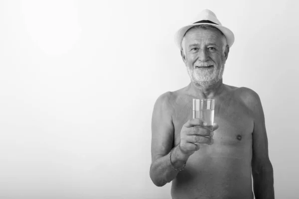 Studio colpo di felice anziano barbuto uomo sorridente mentre tiene il bicchiere d'acqua e indossa cappello senza maglietta sullo sfondo bianco — Foto Stock