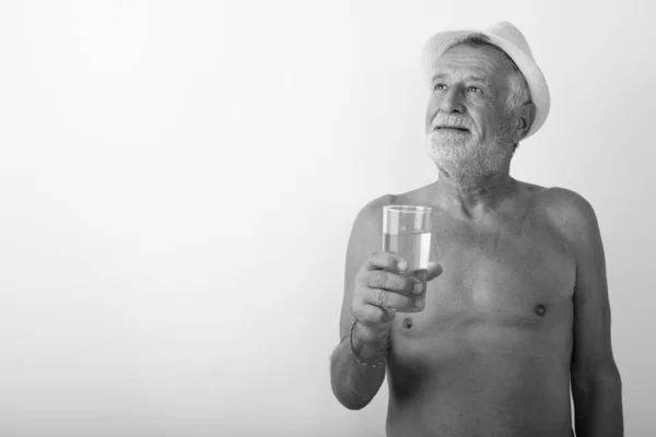 Studio záběr šťastný starší vousatý muž s úsměvem a myšlení při držení sklenice vody s kloboukem bez košile na bílém pozadí — Stock fotografie