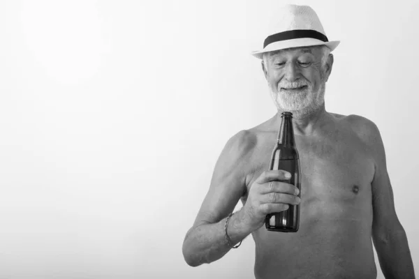 Studio záběr šťastný starší vousatý muž s úsměvem při pohledu na láhev piva a nosí klobouk bez košile na bílém pozadí — Stock fotografie
