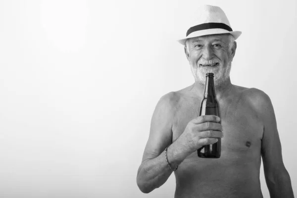Studio colpo di felice anziano barbuto uomo sorridente mentre tiene in mano una bottiglia di birra e indossa cappello senza maglietta su sfondo bianco — Foto Stock