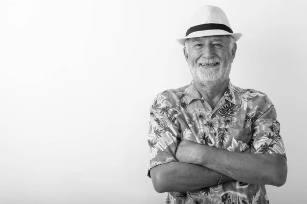 Studio skott av glad senior skäggig turist man ler samtidigt bär hatt med armarna korsade mot vit bakgrund — Stockfoto