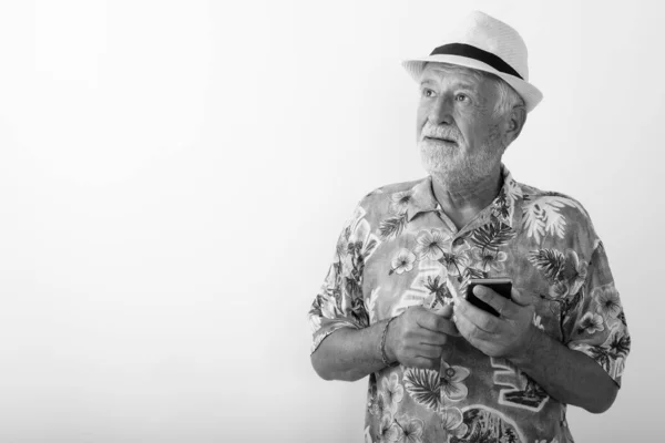 Stúdió lövés jóképű idősebb szakállas turista gondolkodás közben gazdaság mobiltelefon és visel kalap ellen fehér háttér — Stock Fotó