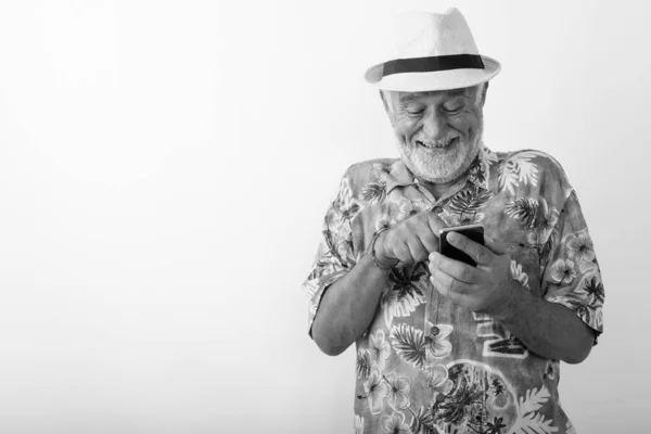 Studio shot van gelukkige senior bebaarde toerist glimlachen en giechelen tijdens het gebruik van mobiele telefoon tegen een witte achtergrond — Stockfoto