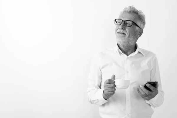 Studio colpo di pensieroso felice uomo barbuto anziano sorridente mentre tenendo tazza di caffè e telefono cellulare con occhiali su sfondo bianco — Foto Stock