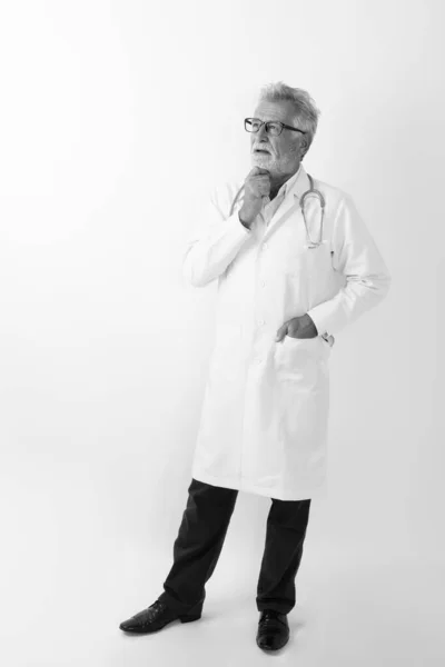 Colpo pieno del corpo di bello anziano barbuto uomo medico in piedi e pensando, mentre guardando sullo sfondo bianco — Foto Stock