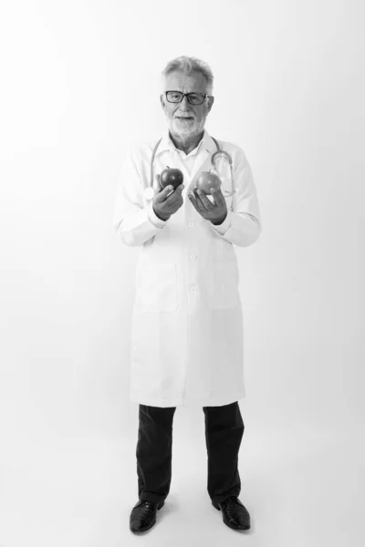 Повний знімок тіла щасливого старшого бородатого лікаря, посміхаючись, тримаючи два яблука на білому тлі — стокове фото