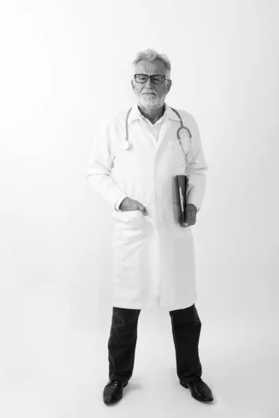 Full body shot di bello anziano barbuto uomo medico in piedi mentre tiene appunti contro sfondo bianco — Foto Stock