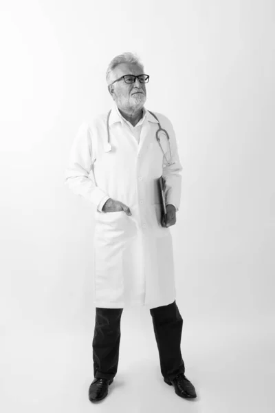 Pieno colpo del corpo di bello anziano barbuto uomo medico in piedi e pensando mentre tenendo appunti sullo sfondo bianco — Foto Stock