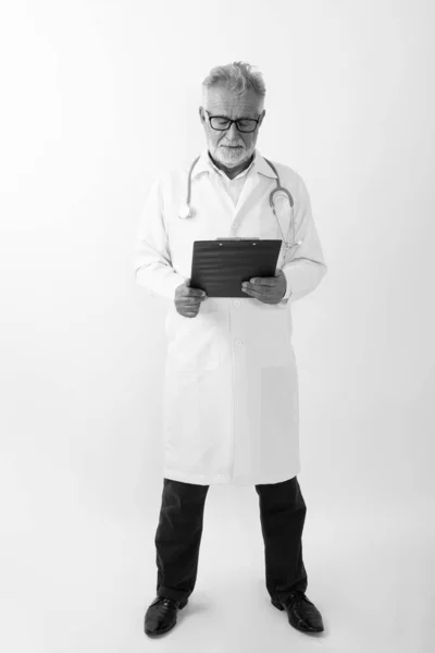Full body shot di bello anziano barbuto uomo medico in piedi durante la lettura sugli appunti contro sfondo bianco — Foto Stock