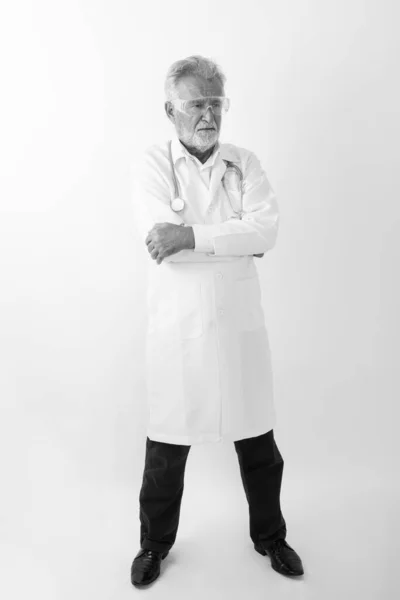 Повний знімок тіла красивого старшого бородатого лікаря, який стоїть під час мислення та носіння захисних окулярів з руками, схрещеними на білому тлі — стокове фото
