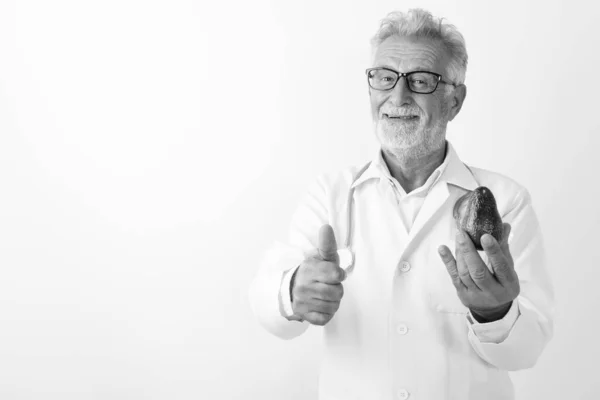Studio colpo di felice anziano barbuto uomo medico sorridente tenendo avocado e dando pollice su sullo sfondo bianco — Foto Stock