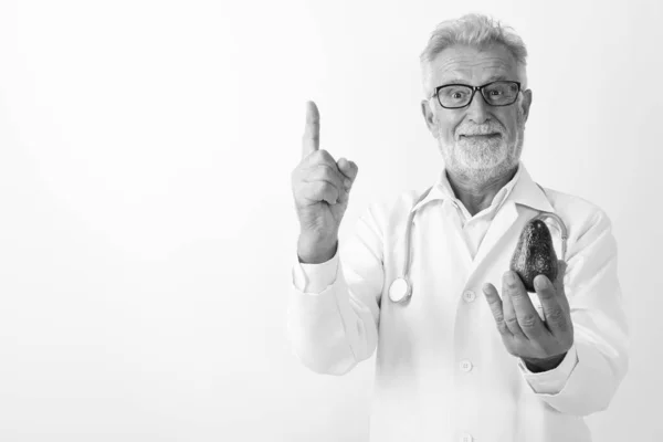 스튜디오는 흰색 배경에 대고 손가락을 가리키면서 아보카도를 들고 있는 건강 한 노인 의사의 사진을 찍었다 — 스톡 사진