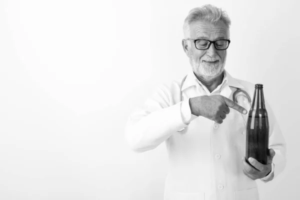 Studio colpo di felice anziano barbuto uomo medico sorridente mentre guardando e indicando bottiglia di birra su sfondo bianco — Foto Stock