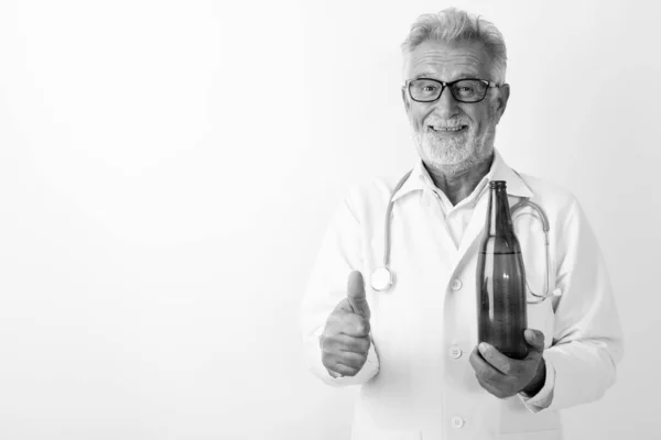 Studio colpo di felice anziano barbuto uomo medico sorridente, mentre tenendo in mano una bottiglia di birra e dando pollice su sfondo bianco — Foto Stock
