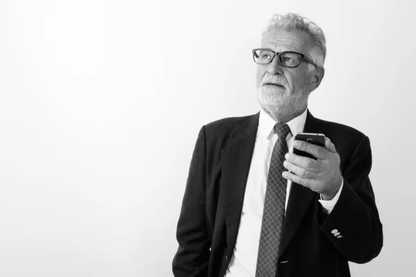 Studio shot van knappe senior bebaarde zakenman denken terwijl het houden van mobiele telefoon tegen een witte achtergrond — Stockfoto