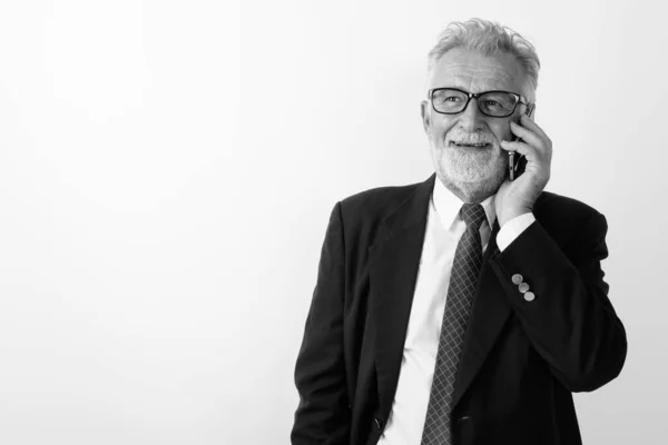 Estúdio tiro de feliz empresário barbudo sênior sorrindo ao pensar e falar no telefone celular contra fundo branco — Fotografia de Stock