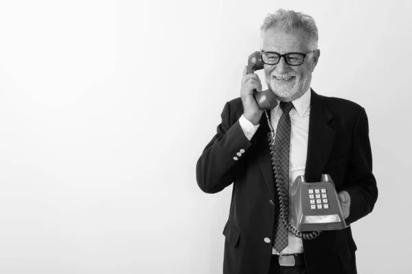 Studio shot van gelukkige senior bebaarde zakenman glimlachend terwijl denken en praten op oude telefoon tegen witte achtergrond — Stockfoto