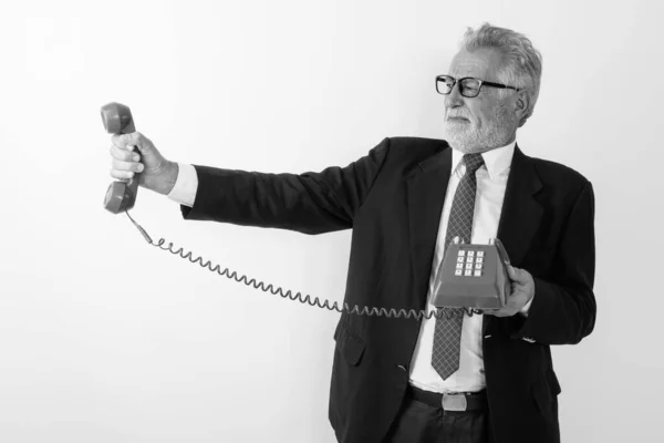 Studio shot van senior bebaarde zakenman houden en kijken naar oude telefoon, terwijl op zoek walgelijk tegen witte achtergrond — Stockfoto