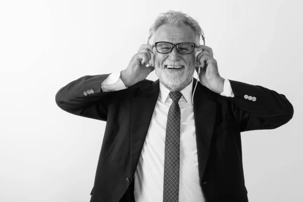 Studio colpo di felice anziano barbuto uomo d'affari sorridente durante l'ascolto di musica e tenendo le cuffie sullo sfondo bianco — Foto Stock
