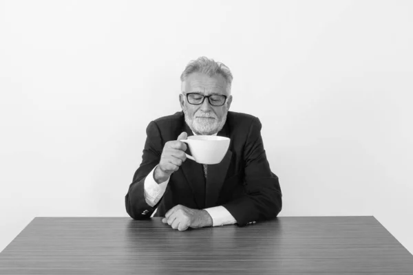 Studio skott av stilig seniorskäggig affärsman som håller och tittar på kaffekopp medan du sitter på träbord mot vit bakgrund — Stockfoto