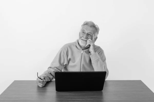 Studio shot szczęśliwego starszego brodatego mężczyzny uśmiechającego się i myślącego trzymając okulary z laptopem na drewnianym stole na białym tle — Zdjęcie stockowe