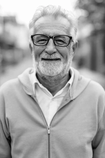 Face of happy handsome senior bearded man smiling while wearing jacket and eyeglasses outdoors