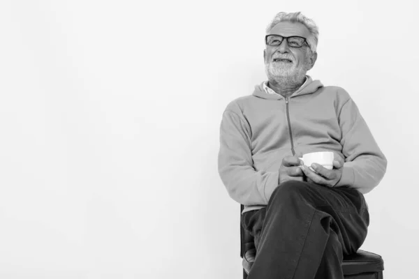 Studio colpo di pensieroso felice uomo barbuto anziano sorridente mentre tiene in mano la tazza di caffè e seduto sulla sedia pronto per la palestra su sfondo bianco — Foto Stock