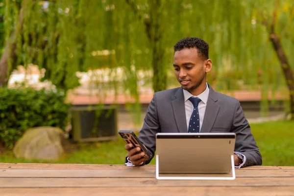 Empresario africano sentado al aire libre usando computadora portátil y teléfono — Foto de Stock