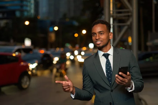 Empresário africano usando aplicativo de telefone celular esperando por táxi com a mão para cima à noite — Fotografia de Stock