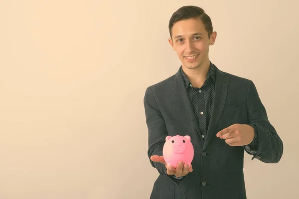 Studio shot van jonge gelukkige zakenman glimlachend terwijl wijzend op piggy bank tegen witte achtergrond — Stockfoto