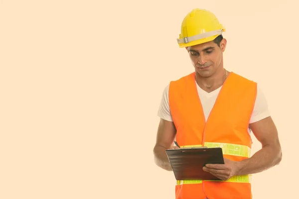 Studio shot of young muscular Persian man construction worker writing on clipboard — Stock Photo, Image