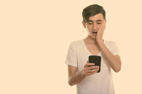 Estúdio tiro de jovem bonito adolescente persa usando telefone celular enquanto olha chocado e cobrindo rosto — Fotografia de Stock