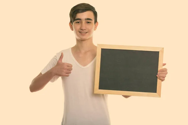 Studio shot van jong gelukkig Perzisch tiener jongen glimlachen terwijl het houden van blanco schoolbord en het geven van duim omhoog — Stockfoto