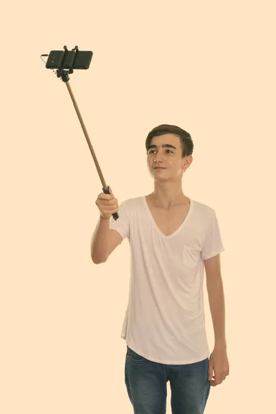 Studio shot of young handsome Persian teenage boy holding selfie stick while taking selfie picture with mobile phone — Stock Photo, Image