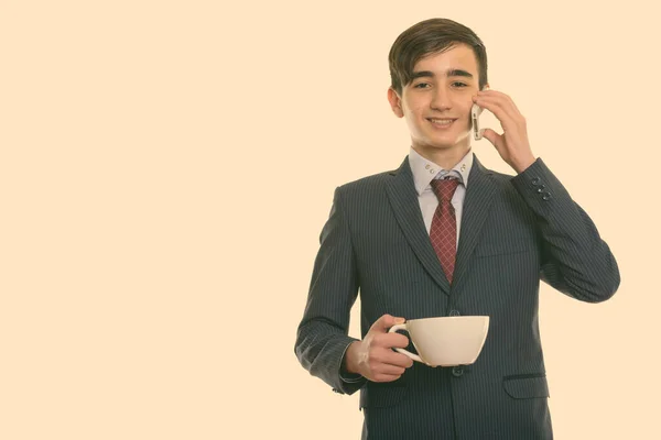 Studioaufnahme eines jungen glücklichen persischen Geschäftsmannes, der lächelt, während er eine Kaffeetasse in der Hand hält und mit dem Handy telefoniert — Stockfoto