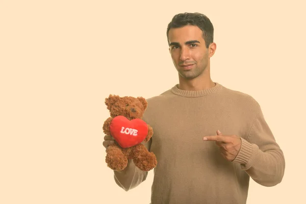 Молодой красивый иранский мужчина держит плюшевого мишку с сердцем и знак любви, указывая пальцем готов к Дню Святого Валентина — стоковое фото