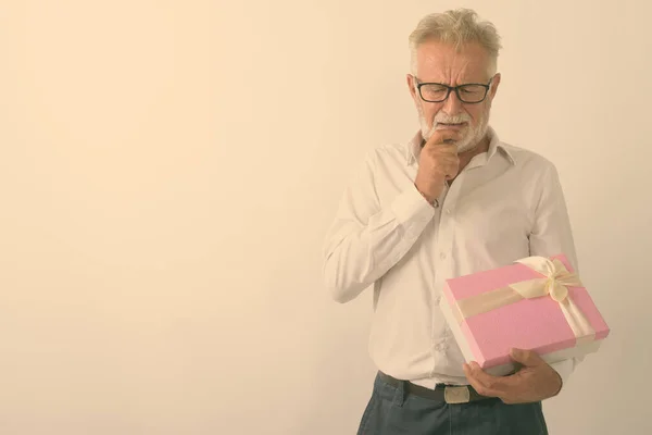 Studio shot of handsome senior bearded man thinking while holding gift box with eyeglasses against white background