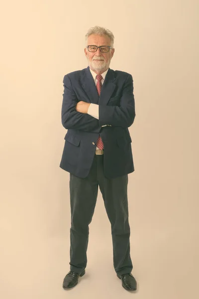 Full body shot of happy senior bearded businessman smiling and standing while wearing eyeglasses with arms crossed against white background