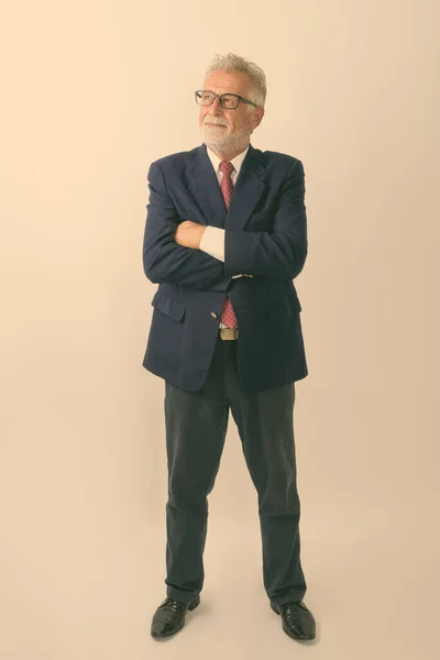 Full body shot of happy senior bearded businessman smiling and standing while thinking with arms crossed against white background