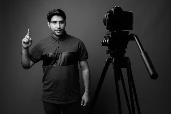 Studio shot of young handsome Iranian man vlogging while wearing gray shirt against brown background