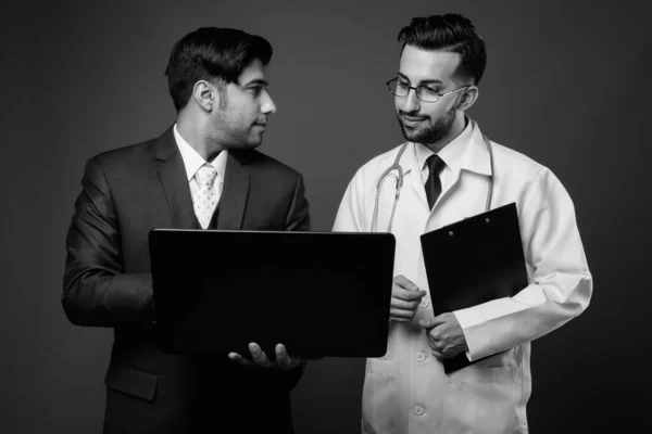 Studio shot of young Iranian businessman and young Iranian man doctor together against brown background