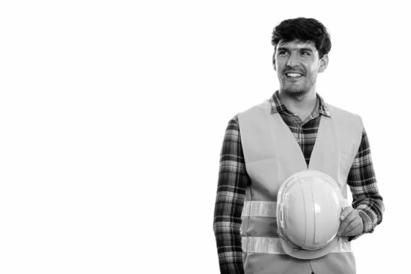 Young happy Persian man construction worker smiling and holding safety helmet while thinking — Stock Photo, Image