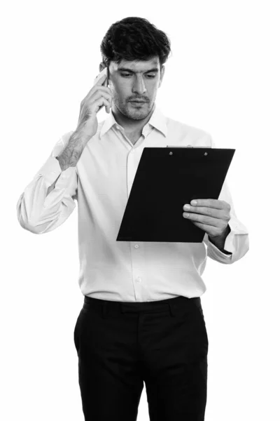 Studio shot van jonge Perzische zakenman lezen op klembord tijdens het praten op mobiele telefoon — Stockfoto