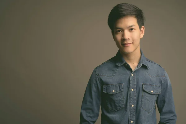 Jonge knappe Aziatische man draagt denim shirt tegen grijze achtergrond — Stockfoto