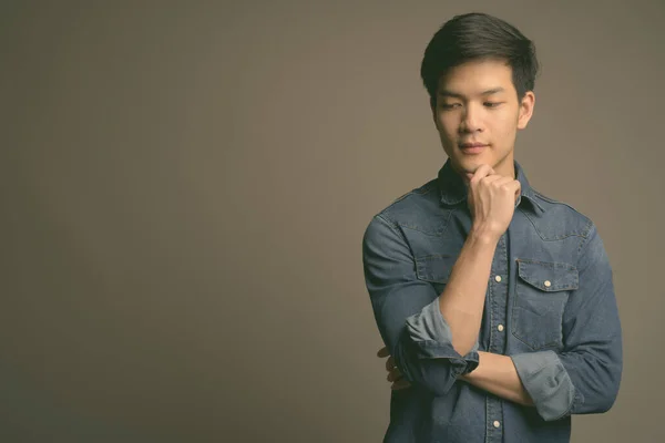 Молодий привабливий азіатський чоловік одягнений у джинсову сорочку на сірому фоні. — стокове фото