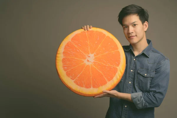 Joven guapo asiático hombre sosteniendo naranja almohada sobre gris fondo — Foto de Stock