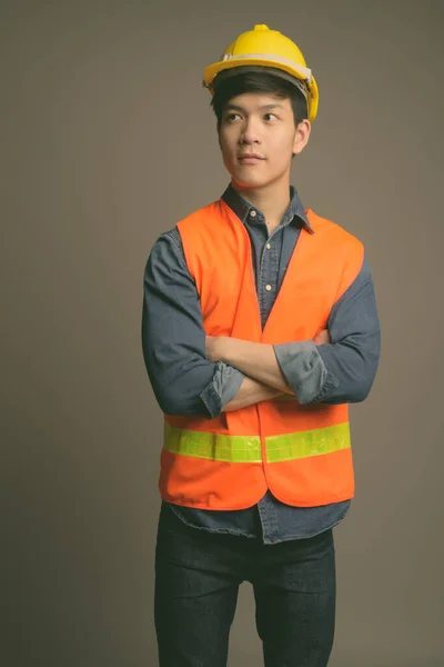 Junger hübscher asiatischer Mann Bauarbeiter vor grauem Hintergrund — Stockfoto